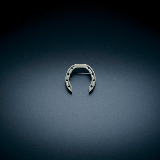 A sleek horseshoe-shaped brooch without stones, displayed on a dark, textured background. The brooch features a minimalist design with a horizontal clasp on the back.