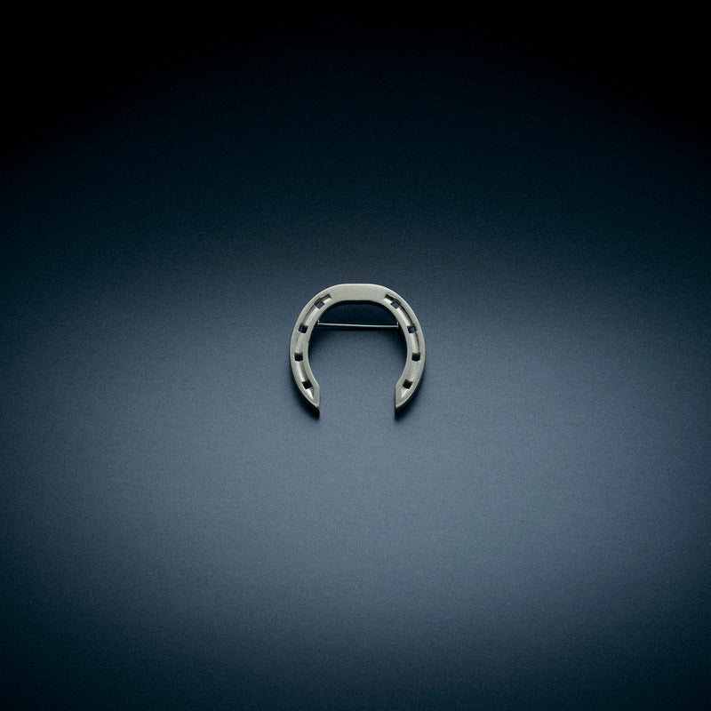 A sleek horseshoe-shaped brooch without stones, displayed on a dark, textured background. The brooch features a minimalist design with a horizontal clasp on the back.