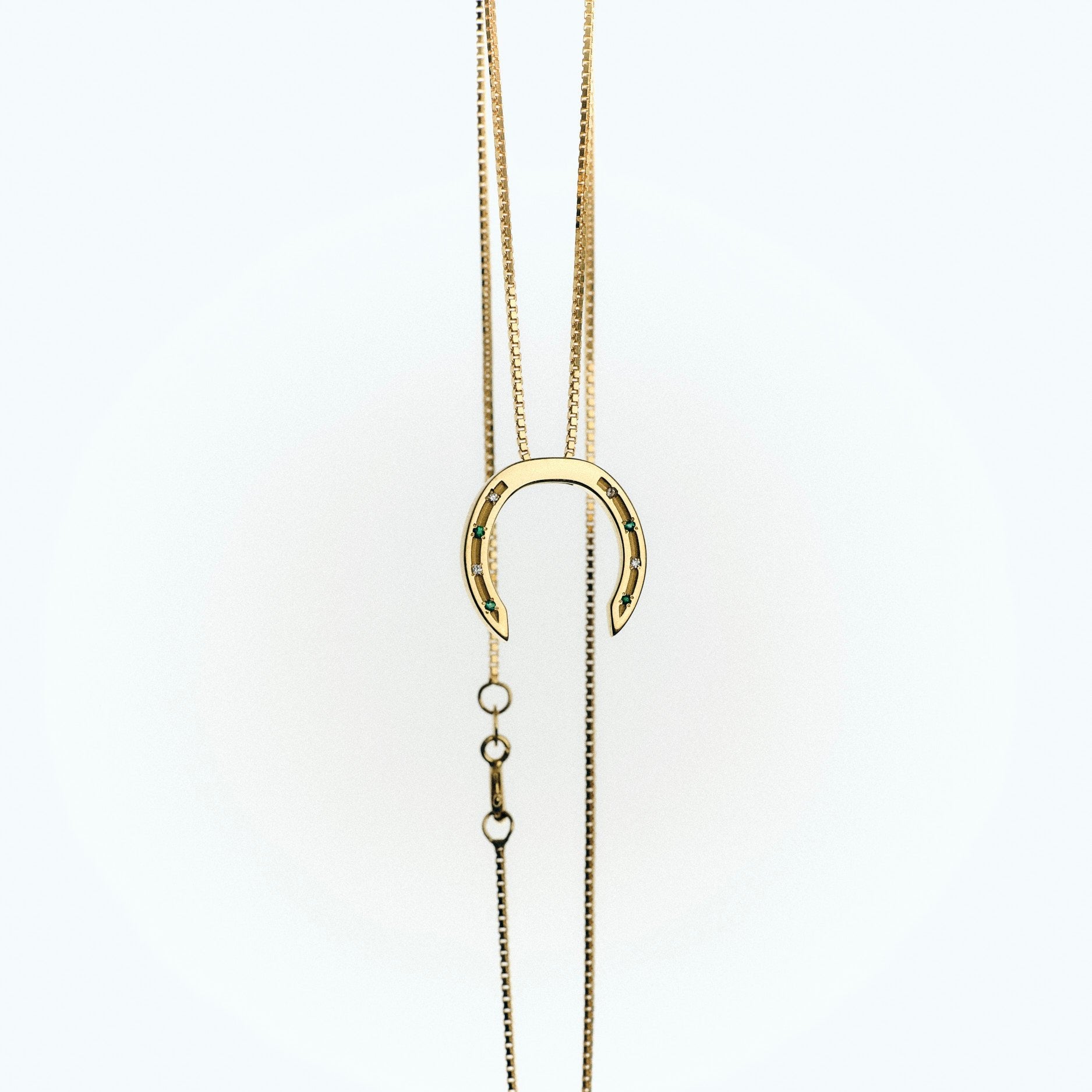 A delicate gold plated necklace with a horseshoe pendant, featuring small diamonds and emerald stones embedded in the design. The chain is elegantly draped, with a clasp visible at the bottom, set against a light background.